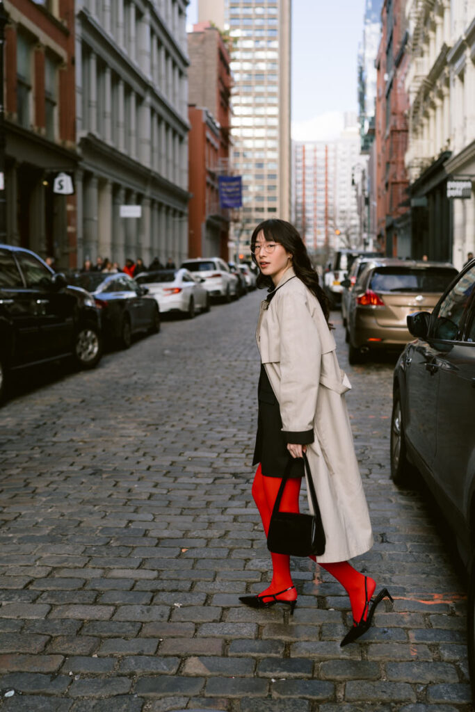 A fashionable person walking on a cobblestone road, turned to look over their shoulder, wearing red tights, black heels, and a beige coat, with a black Gucci bag in hand