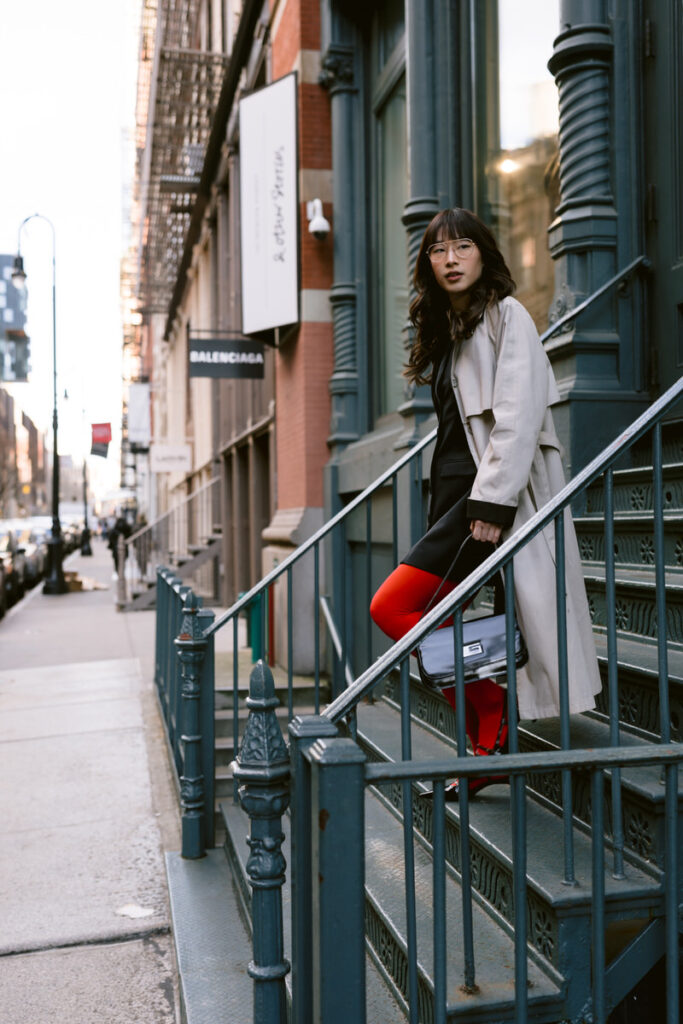 A stylish person posing on city stairs, wearing red tights, a black dress, and a beige trench coat, holding a black Gucci bag, with a Balenciaga sign in the background