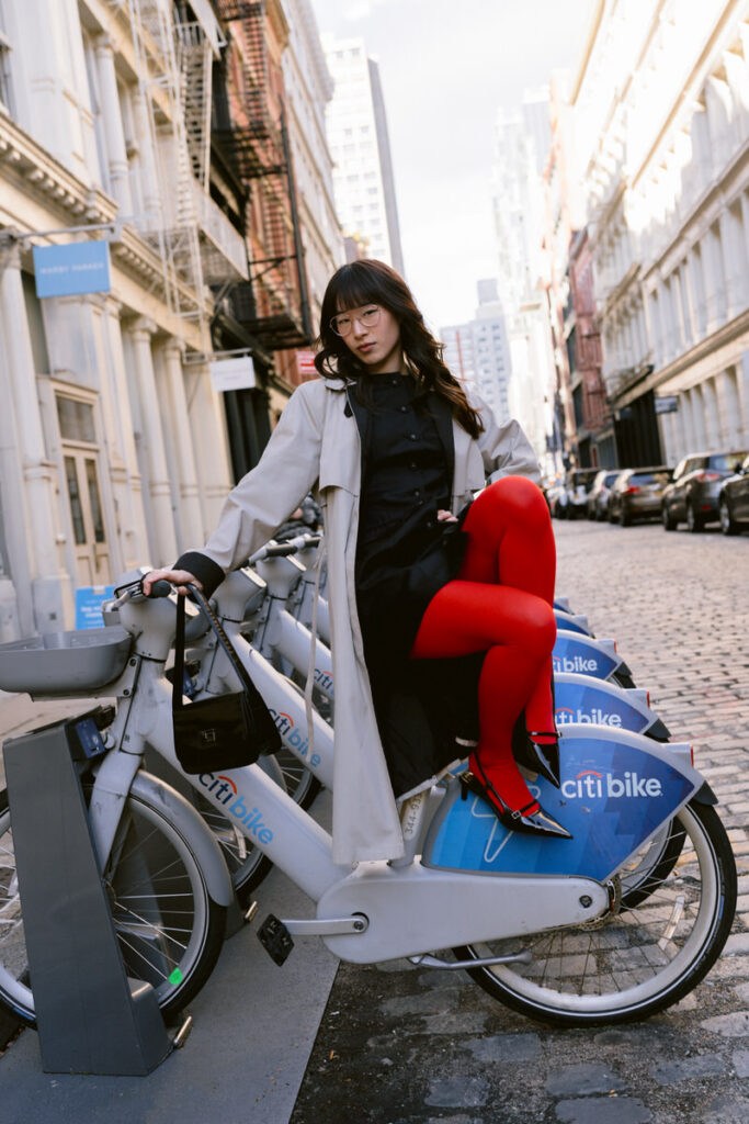 A chic individual seated on a blue Citibike, sporting red tights and a black dress layered under a beige coat, complemented by a black Gucci bag and stylish glasses