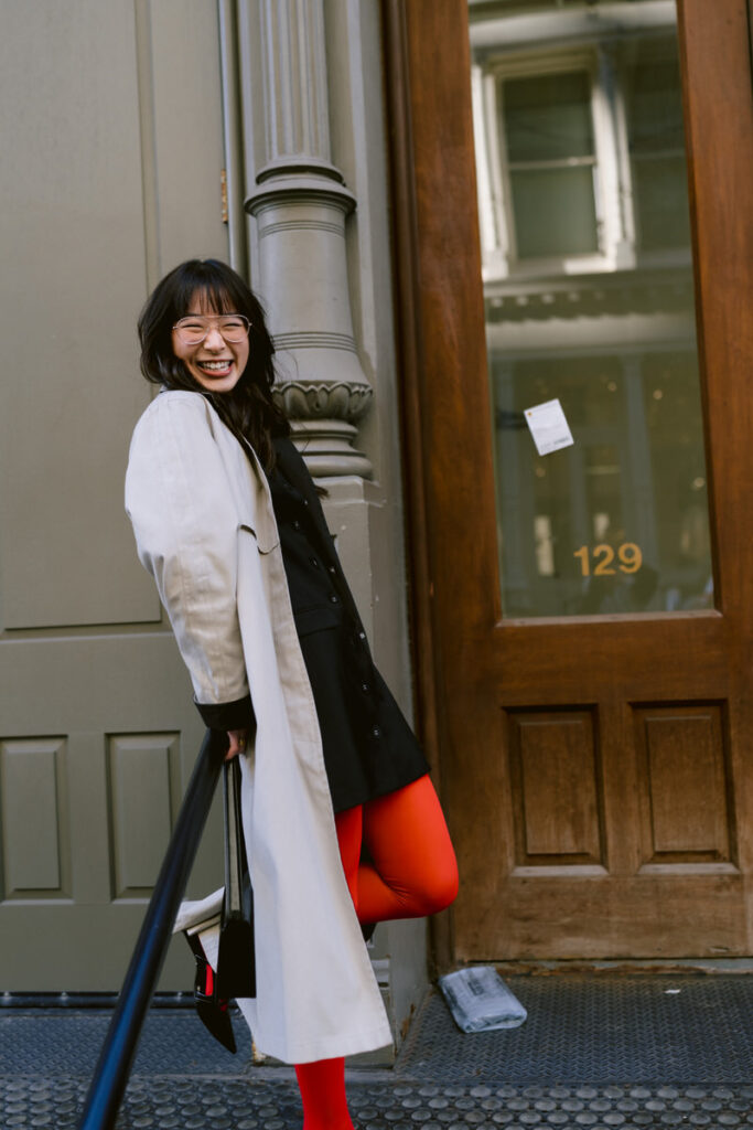 A joyful person standing by a doorway, laughing and clutching a black Gucci bag, dressed in a black dress, red tights, and a casual beige coat