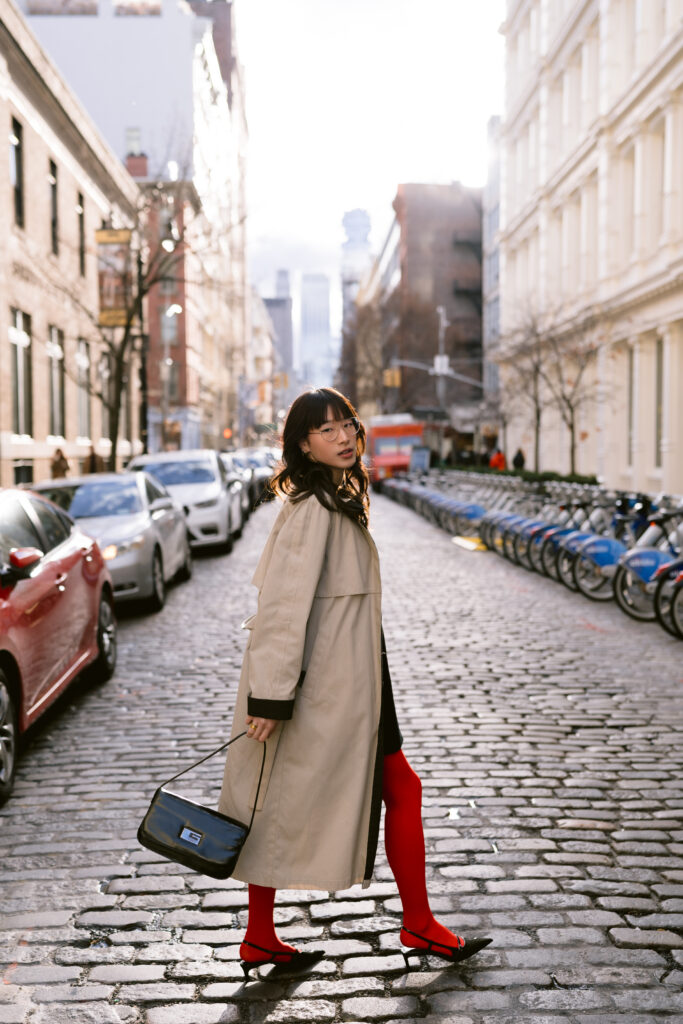 A thoughtful individual standing on a cobblestone street, gazing into the distance, dressed in a beige trench coat, black dress, and striking red tights, holding a black Gucci bag