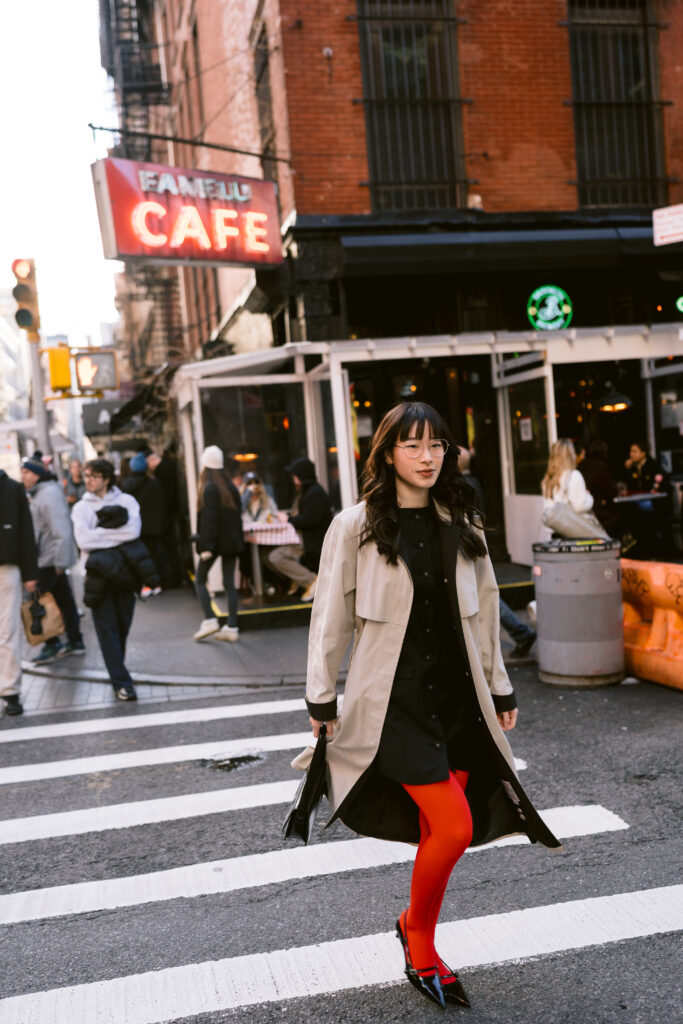 A trendy individual walking across a a crosswalk in a bustling cityscape, sporting a beige coat, black dress, and vibrant red tights, with a black Gucci bag in hand