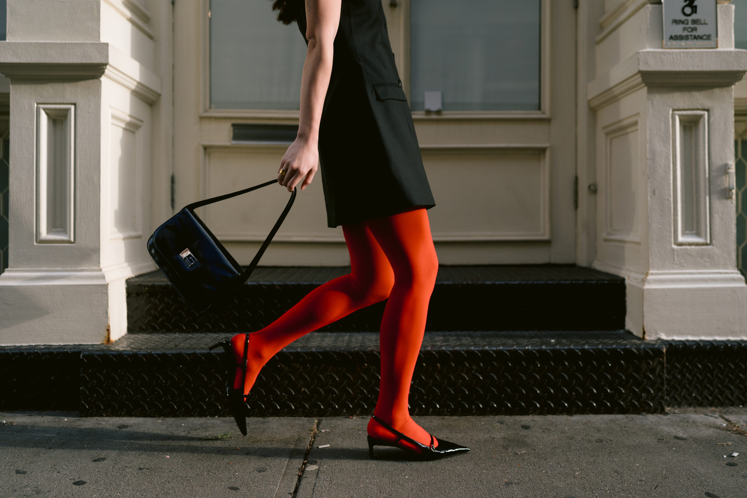 Content creation photography featuring a stylish stride captured mid-movement, featuring a person in red tights and black slingback heels, carrying a black Gucci bag, against an urban backdrop with white architectural details