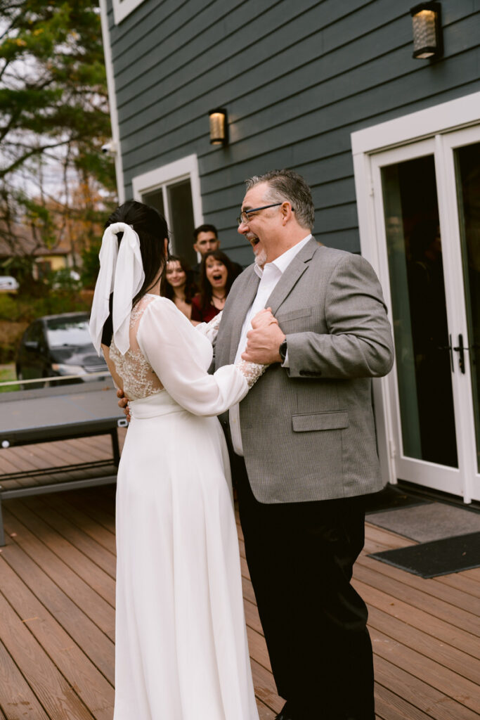 Laughing bride hugging her father, on a wooden deck with people cheering in the background.
