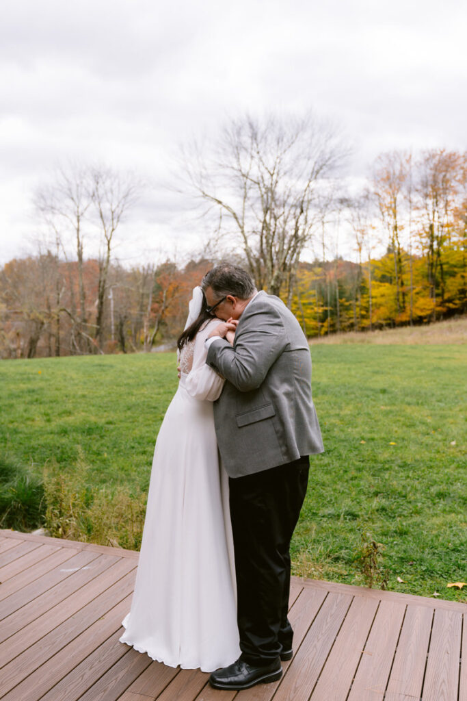 A tender moment of a bride and her father embracing on a wooden deck with a backdrop of fall trees and overcast sky.
