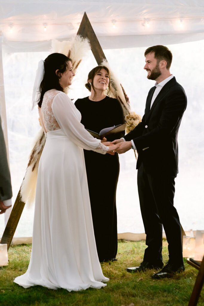 A bride and groom holding hands, exchanging vows in front of an officiant, under a wedding tent with a bohemian triangular arch decorated with pampas grass.
