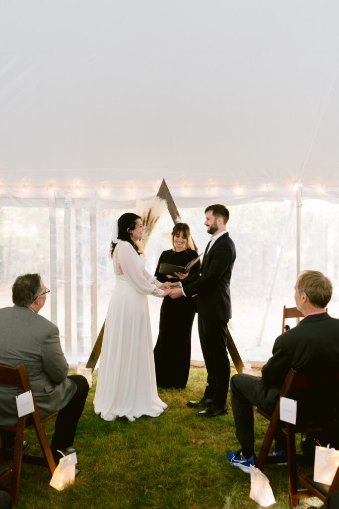 A bride and groom exchanging rings during their wedding ceremony under a tent, with a geometric wooden arch and onlooking guests.

