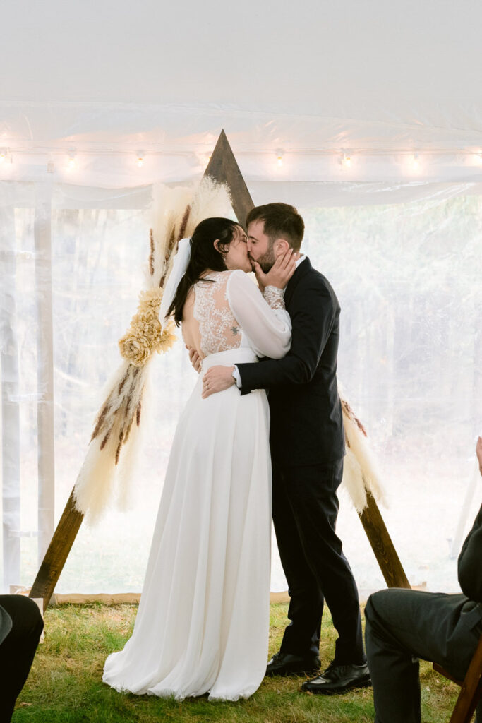 A bride and groom sharing their first kiss as a married couple under a tented altar with a rustic wooden triangle arch and feathery pampas grass.
