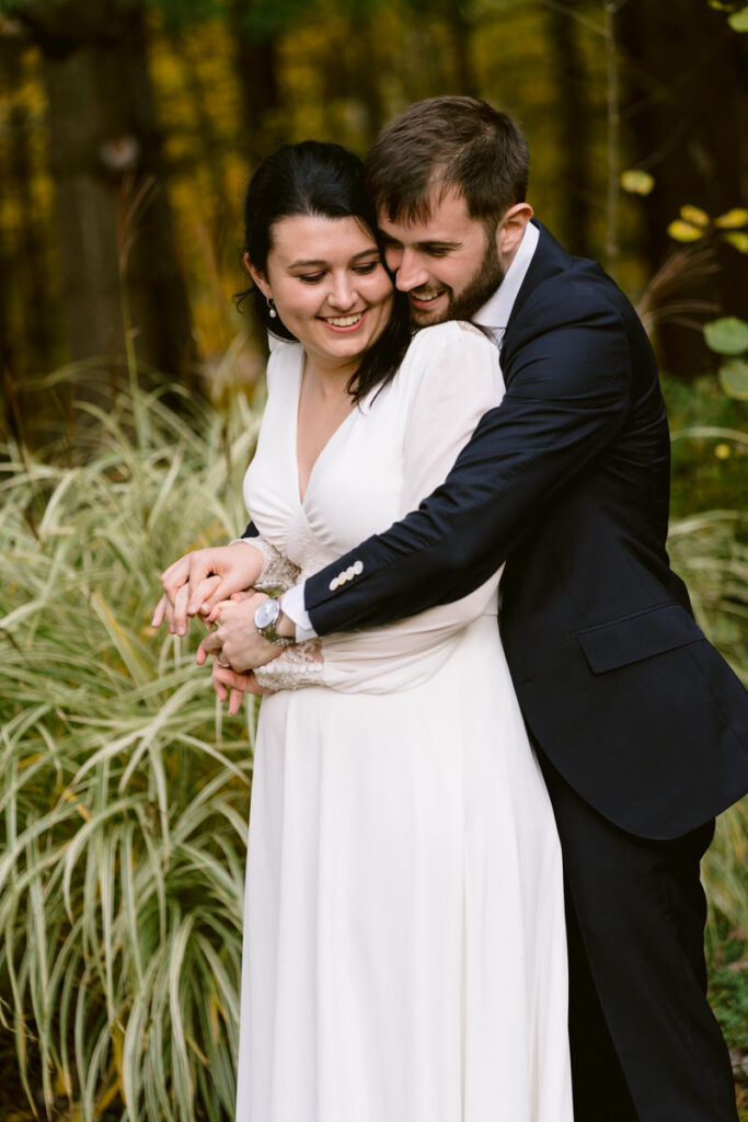An intimate moment between a bride and groom embracing each other, with the bride looking over her shoulder, surrounded by lush greenery.
