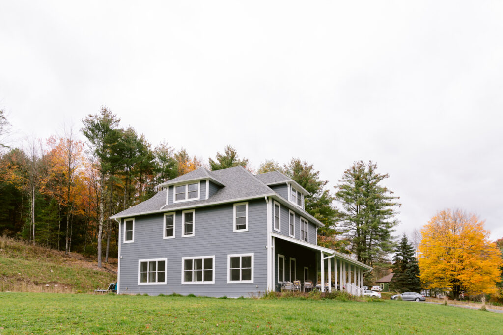 A large gray house with a wrap-around porch in a rural setting, surrounded by trees with autumn leaves.
