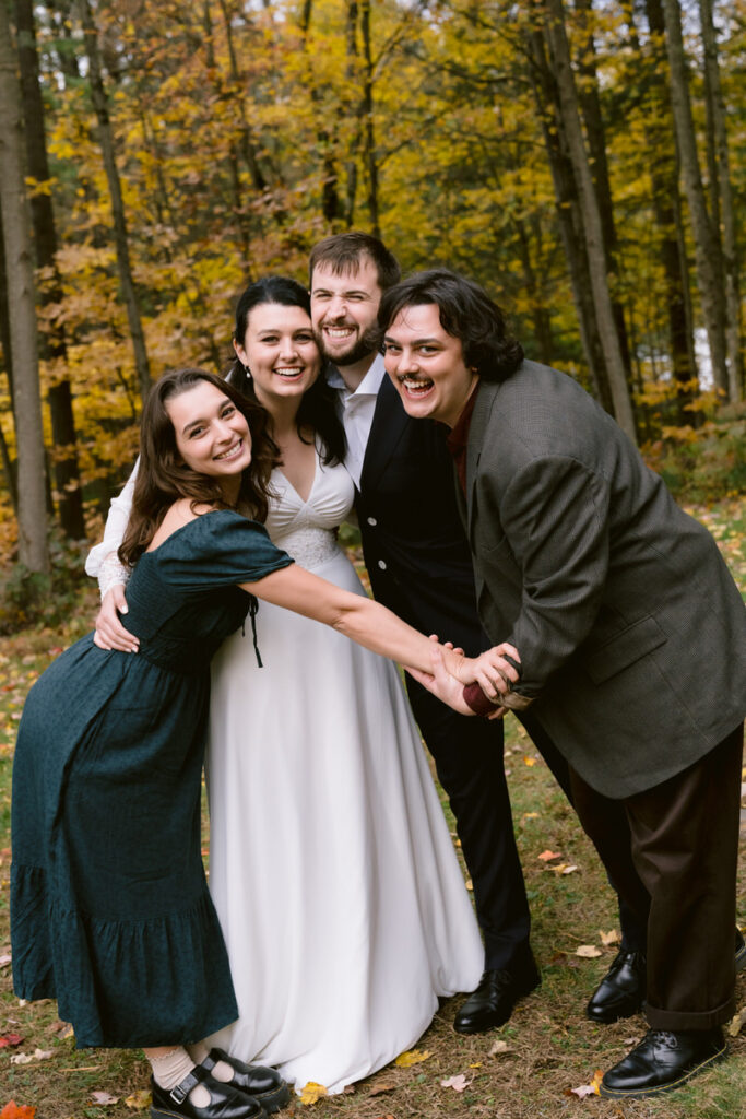 A group photo of a bride, groom, and two guests smiling and holding hands, with a forest with autumn foliage in the background.

