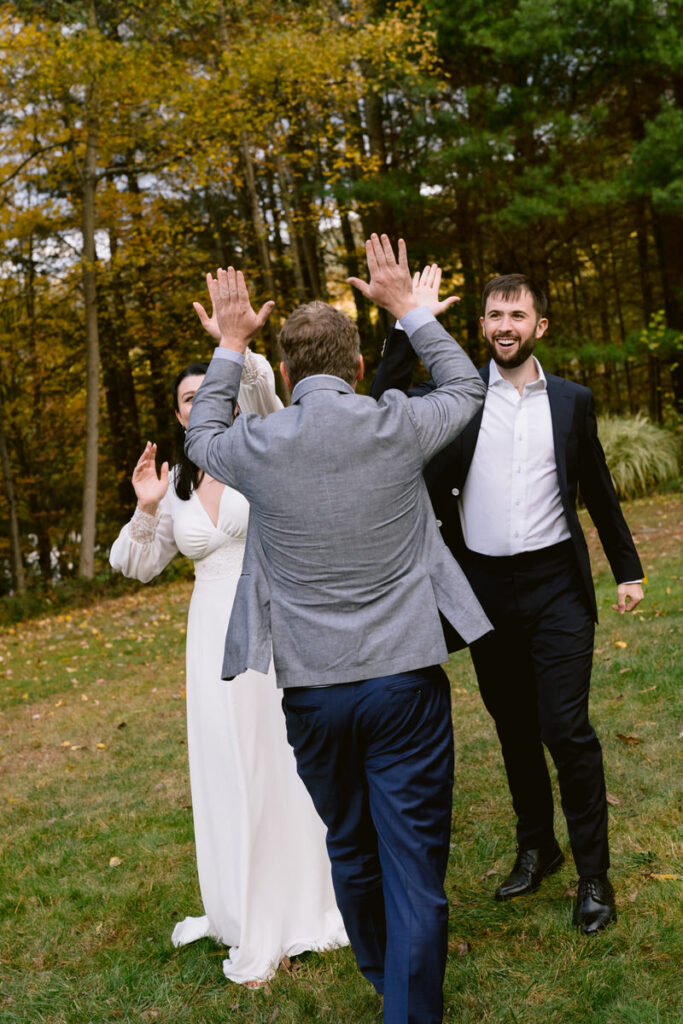 Joyful bride and groom high-fiving a guest in a casual outdoor wedding setting with autumn trees in the background.
