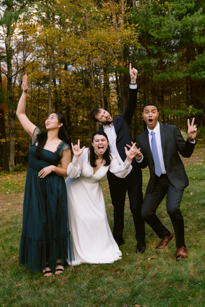 Wedding group photo with the bride and groom in the center, surrounded by four guests making playful gestures, with autumn trees in the background.
