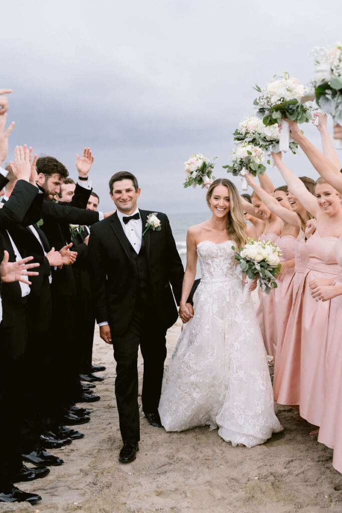 Bride and groom walking hand in hand on the beach, surrounded by bridesmaids in pink dresses and groomsmen in black suits raising their hands in celebration.