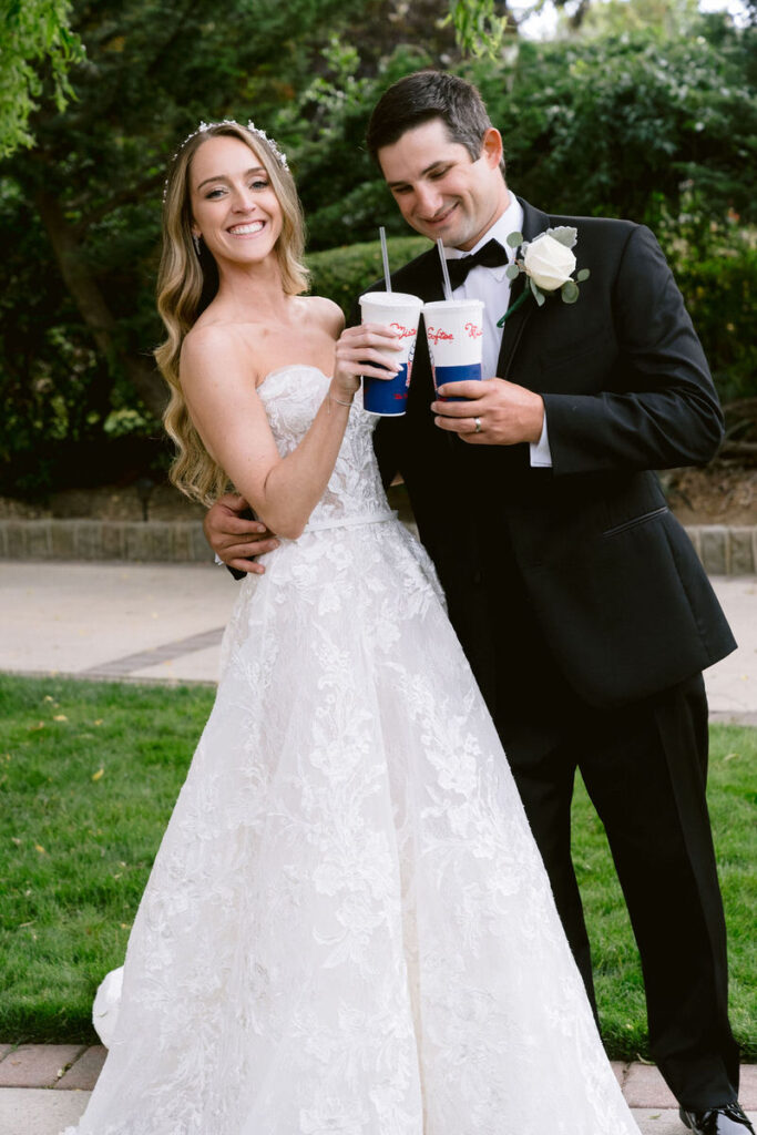 Bride and groom smiling while holding fast food drinks with the logo 'Dutch Bros' on them in a garden setting