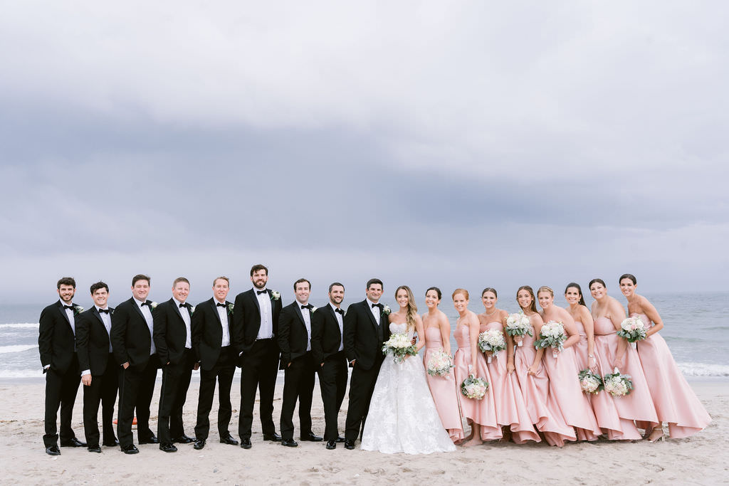 Full bridal party on the beach, with bridesmaids in pink dresses and groomsmen in black suits.