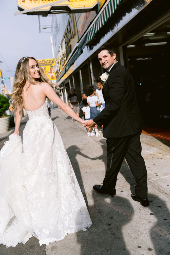 Bride looking back, smiling, as she holds hands with the groom, with a bustling street scene behind them.