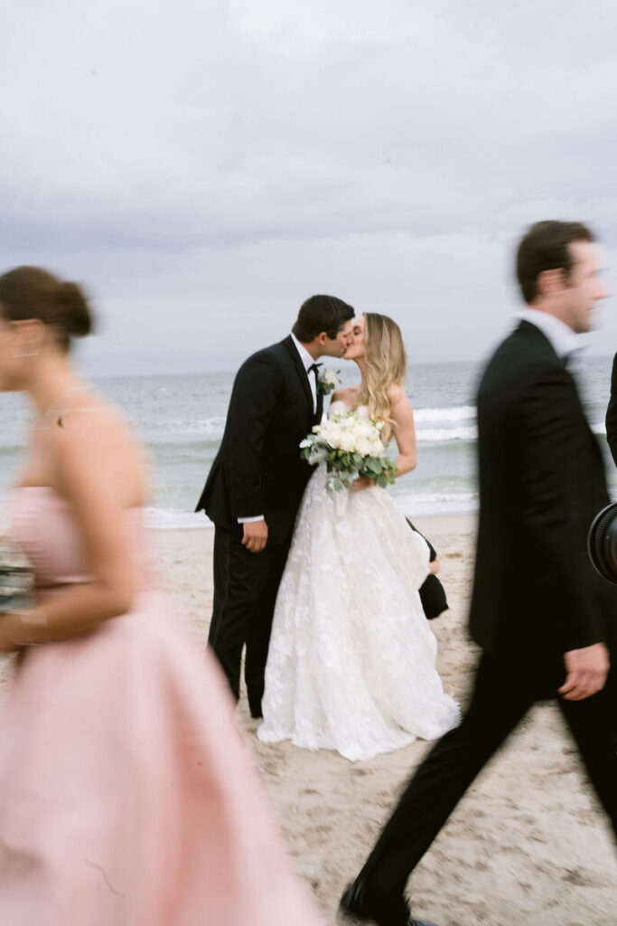 Bride and groom kissing on the beach as guests walk by, capturing a candid moment.