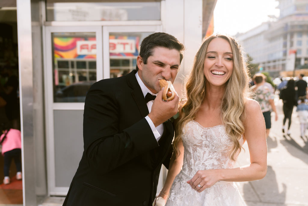 Groom biting into a hotdog with bride laughing beside him, outside a food stall on a sunny day.