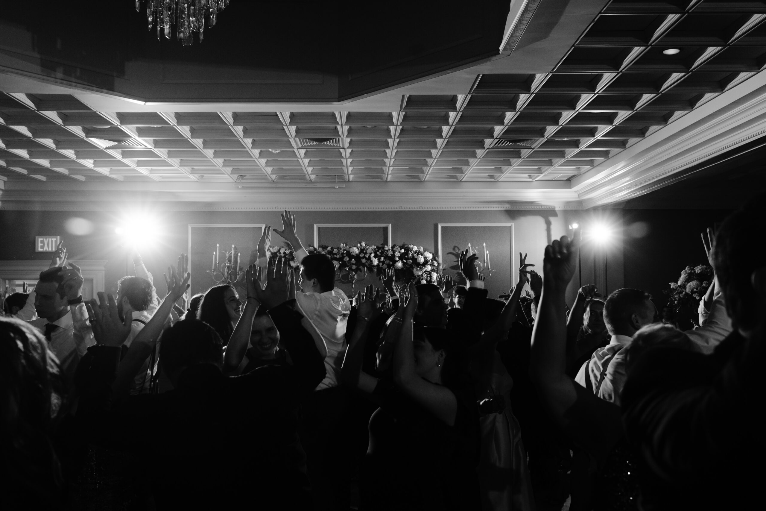 black and white edit of a group of people dancing with their hands in the air. Spotlights are shining behind them.