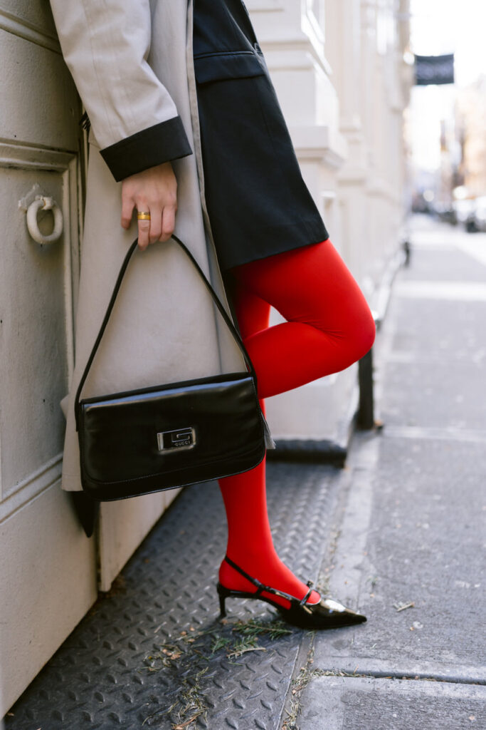 A close-up of red tights and black pointed heels with ankle straps, the subject's hand resting against a cream trench coat, set against an urban street backdrop.