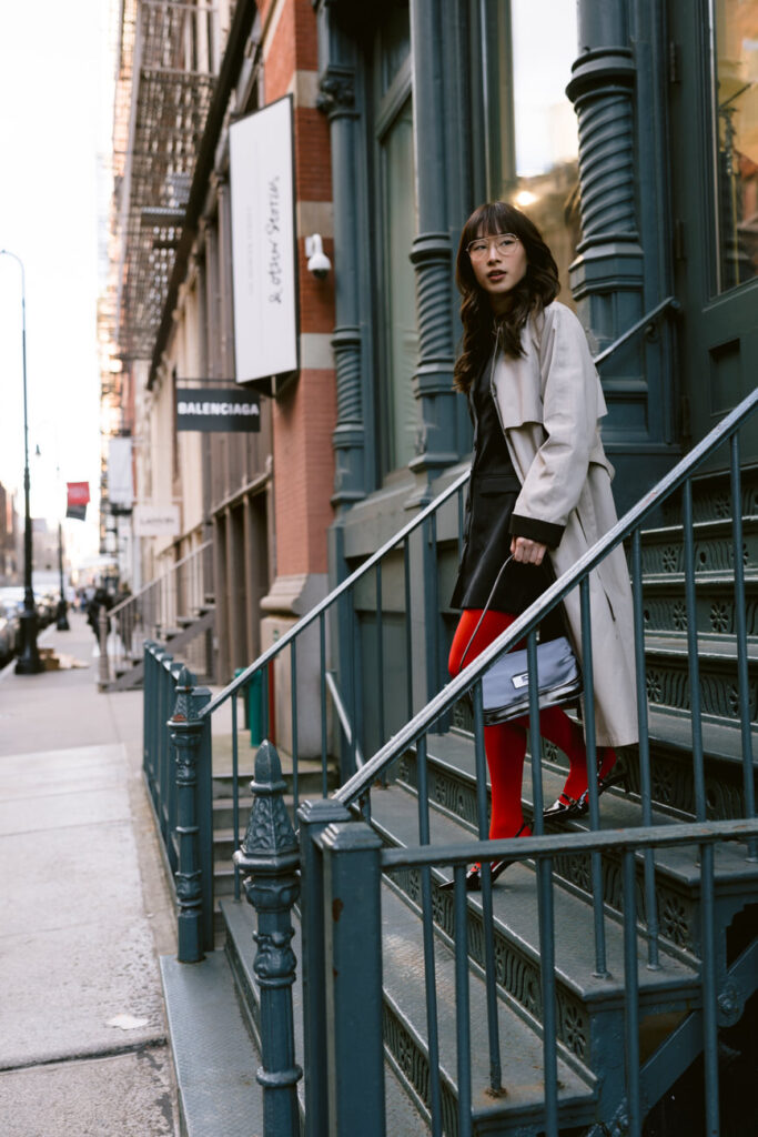 Casually leaning on a metal railing, the subject is styled with a mix of sophistication and edge, sporting a long trench coat, black outfit, red tights, and black boots, with the classic architecture of NYC's West Village in the background.