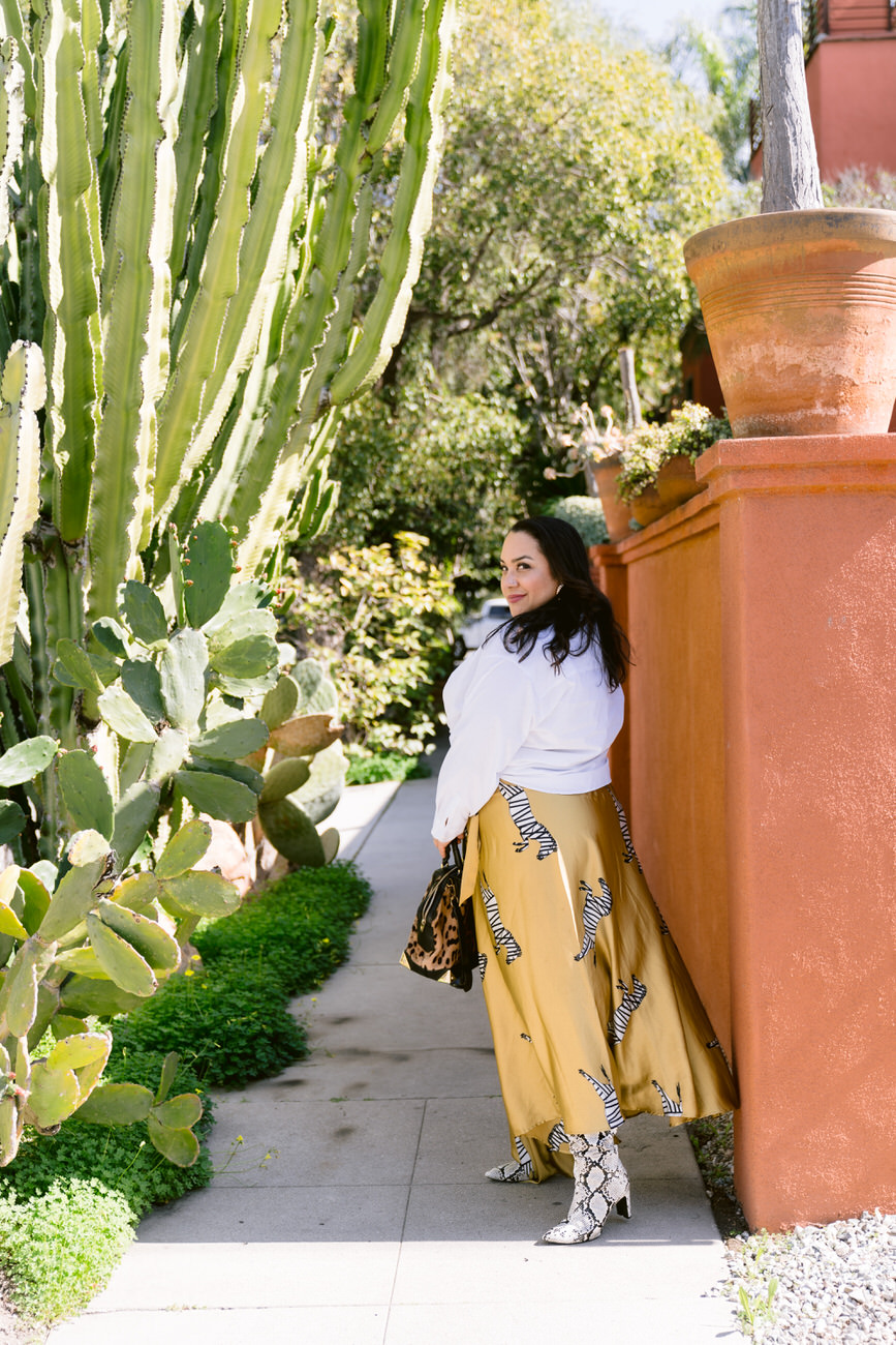 A woman in a stylish white blouse and yellow skirt with a zebra print walks past a tall cactus garden, complemented by her snake print boots and a leopard print bag, exuding a chic vibe in a lush outdoor setting.