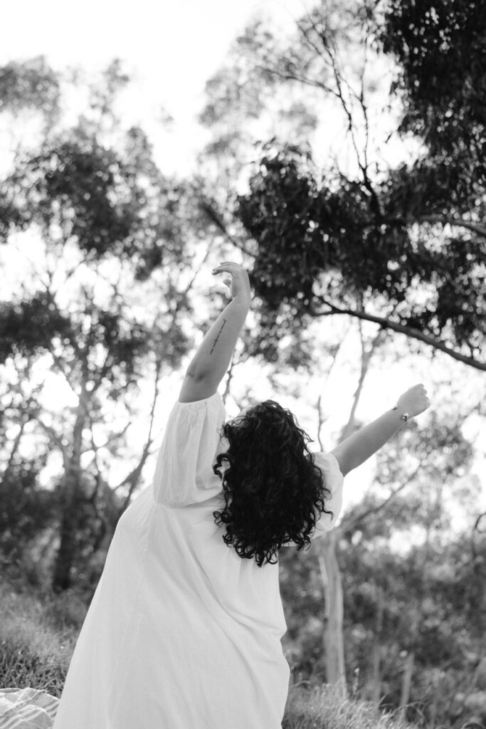 In a black and white photo, a woman in a flowing white dress stands in a field, her arms joyfully raised towards a canopy of trees, capturing a moment of carefree bliss.