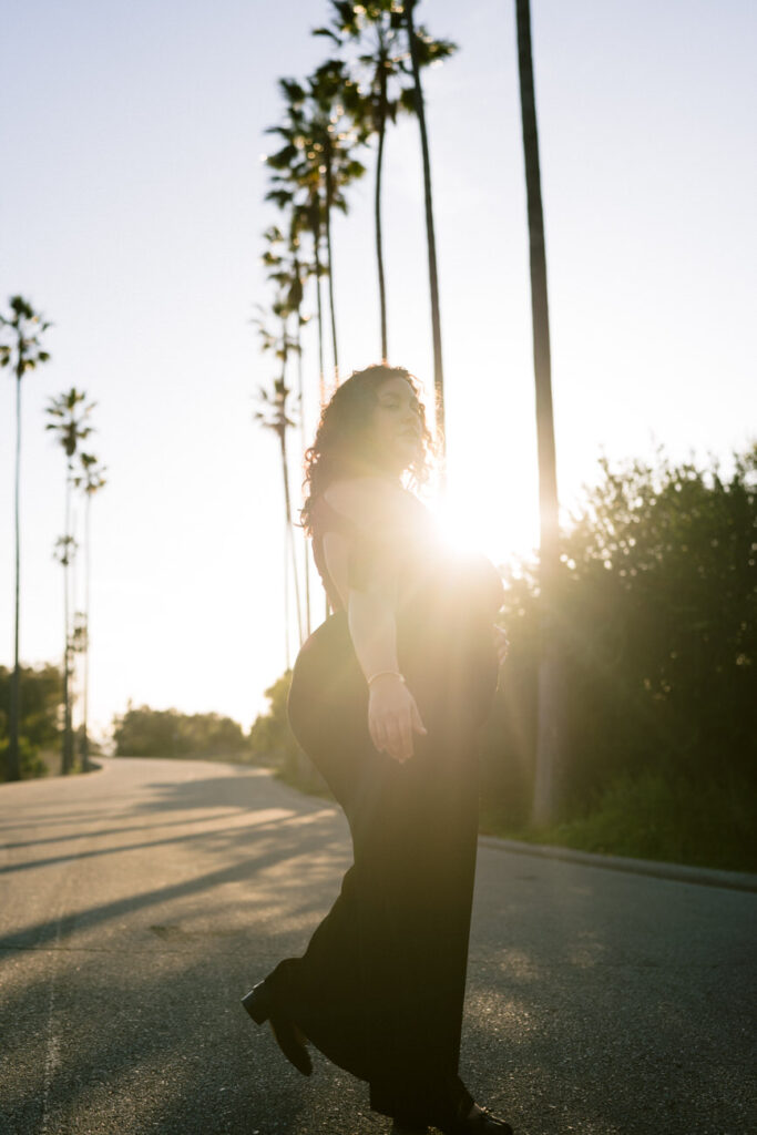 Against the light of a setting sun, a woman in a dark dress walks down a street lined with palm trees, her silhouette illuminated from behind, evoking a serene end-of-day atmosphere.