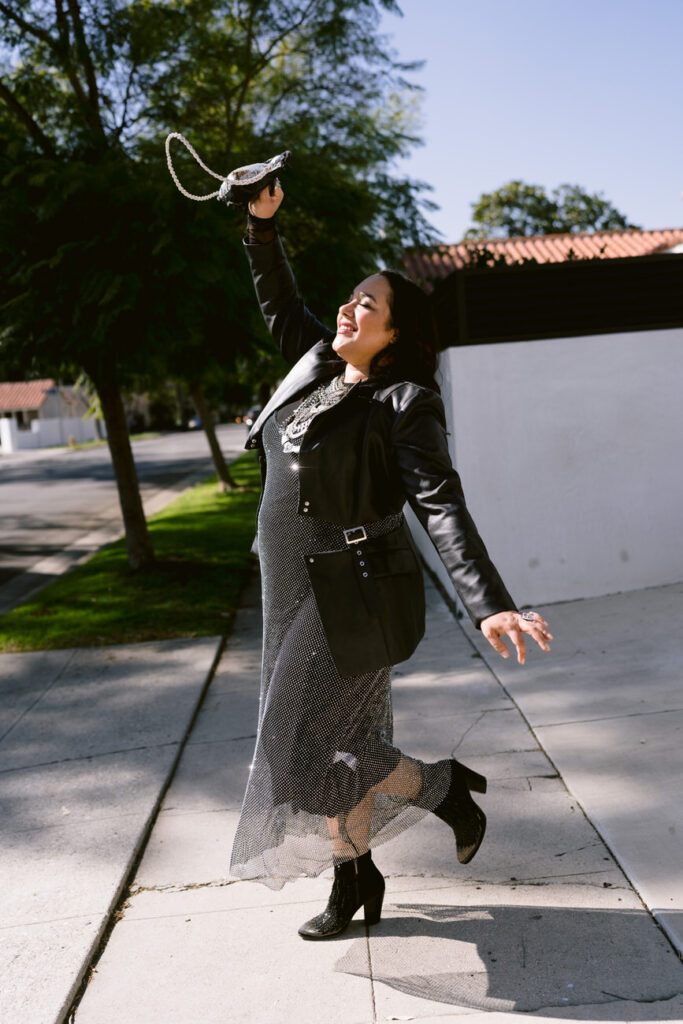 A joyful woman twirls with a snakeskin handbag in hand, dressed in a black leather jacket and a mesh overlay skirt, with textured black boots, reveling in the sunny outdoors.