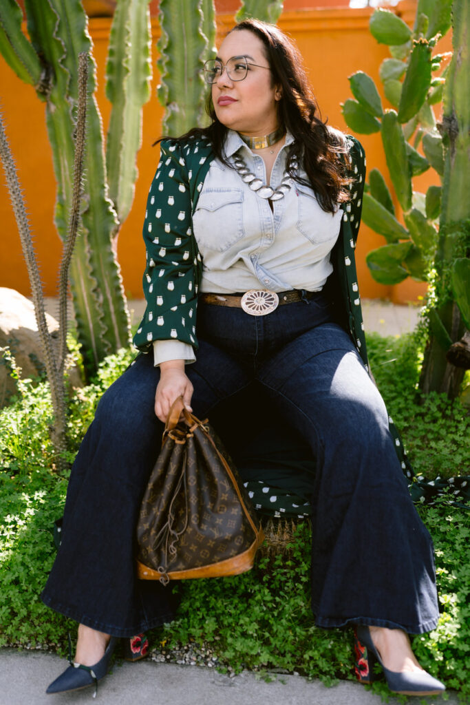 Seated casually, a stylish woman with a polka-dot blouse and denim overalls holds a classic designer drawstring bag, surrounded by a vibrant cactus garden.
