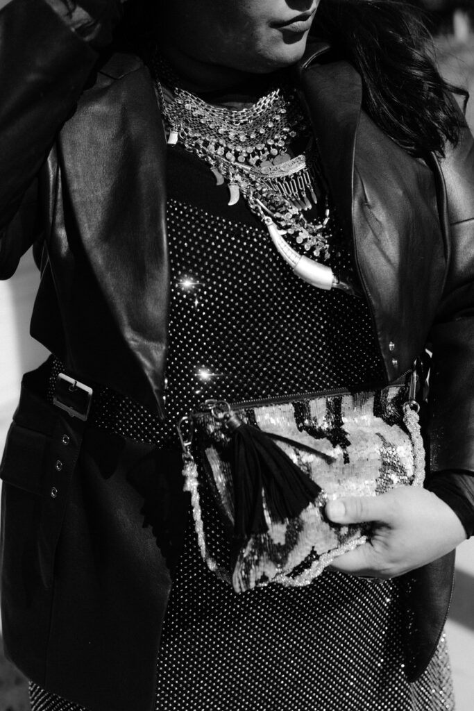 Black and white image focusing on the upper body of a person adorned with a statement silver necklace, wearing a black leather jacket, and holding a sequined clutch bag.