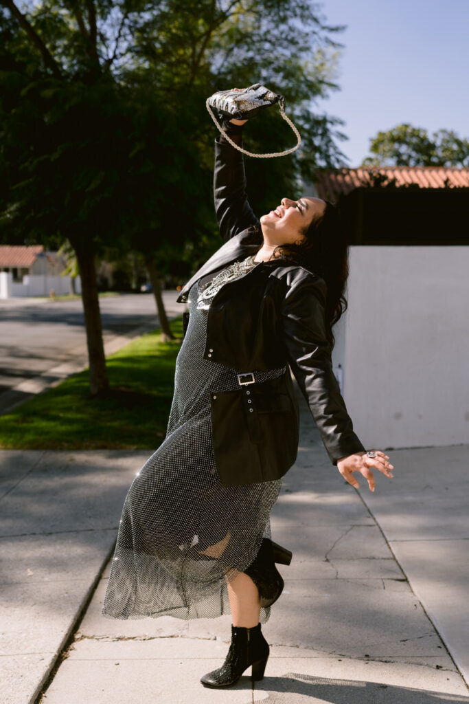 A person joyfully throwing a sequined clutch bag in the air, wearing a black leather jacket and a dotted dress with a glittery sheer overlay, against a bright outdoor background.