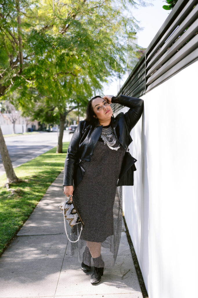 A person leaning against a white wall, looking relaxed, in a black leather jacket and dotted dress with a sparkling sheer layer, clutching a sequined handbag, with trees and sunlight in the background.