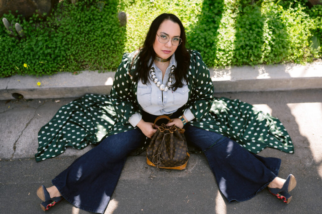 A person sitting on a curb under the shade of a tree, dressed in denim bell-bottoms and a green polka dot kimono, holding a vintage designer bucket bag.