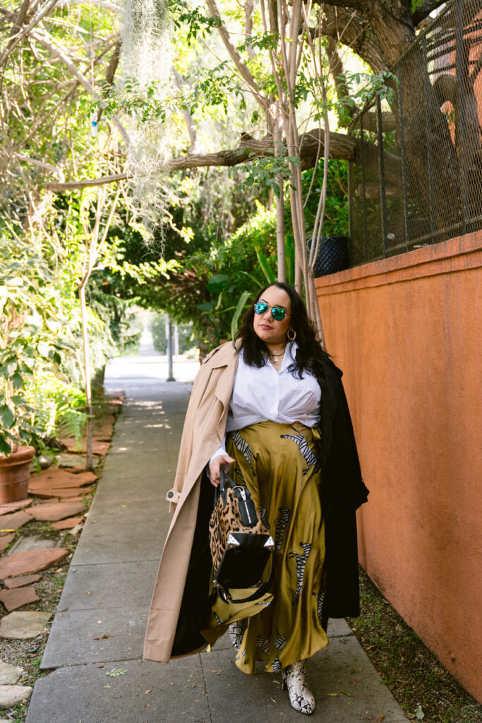A full-body image of a person in a stylish outfit, featuring a white blouse, a mustard skirt with a zebra print, snake print boots, and a leopard print bag, standing on a walkway with lush greenery around.