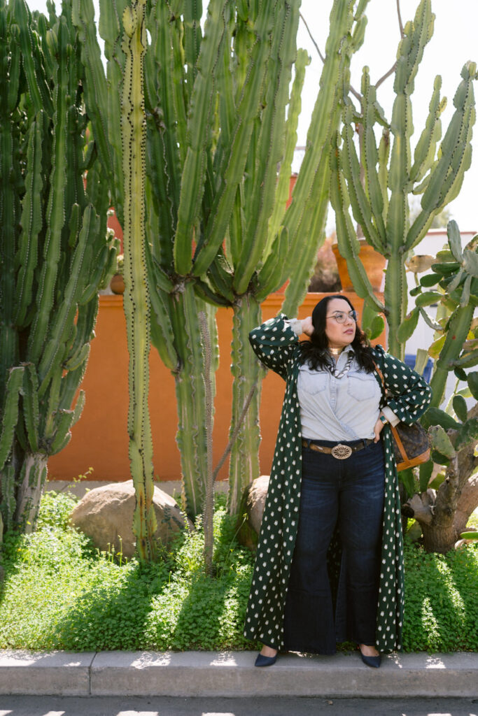 A person posed with hands on hips among tall cacti, wearing a green polka dot kimono over a denim shirt and jeans, complemented by a vintage designer bag.