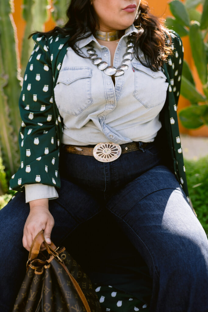 Close-up of a person seated, showcasing a denim shirt, dark jeans, a wide belt with a large round buckle, and a green polka dot kimono, with a designer handbag in hand.