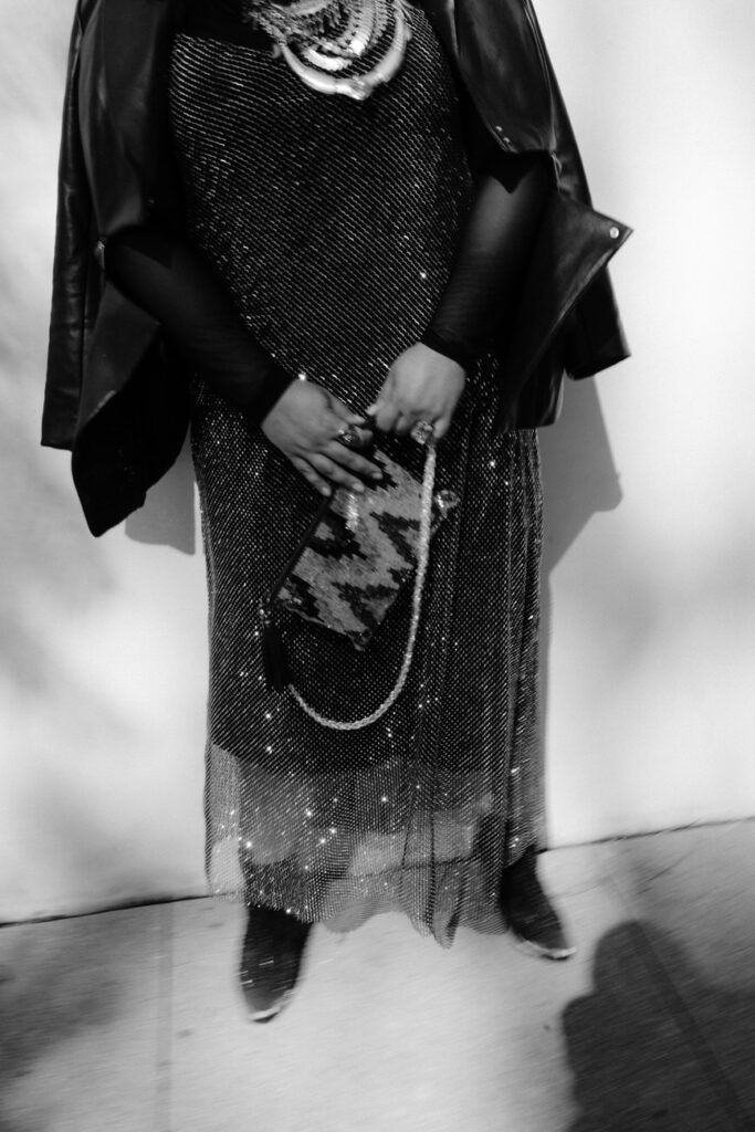A monochrome portrait capturing the joy of a woman in a sequined dress and leather jacket, holding a patterned clutch, her jewelry gleaming as she stands against an urban backdrop.