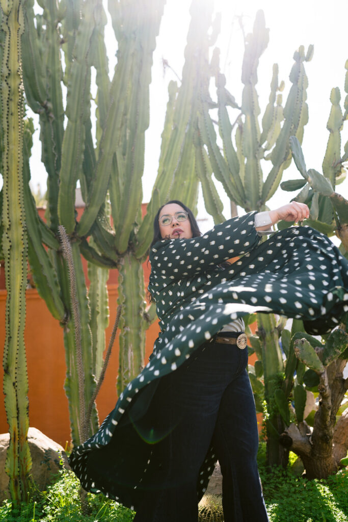 A woman in a green polka-dot blouse throws her arms wide in a whimsical gesture, with a cactus garden behind her, symbolizing freedom and style.