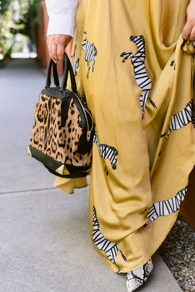 Close-up of a woman's side, holding a leopard print bag, wearing a white blouse and mustard zebra-striped skirt, complemented by snake print boots, focusing on the dynamic textures and patterns of her outfit.