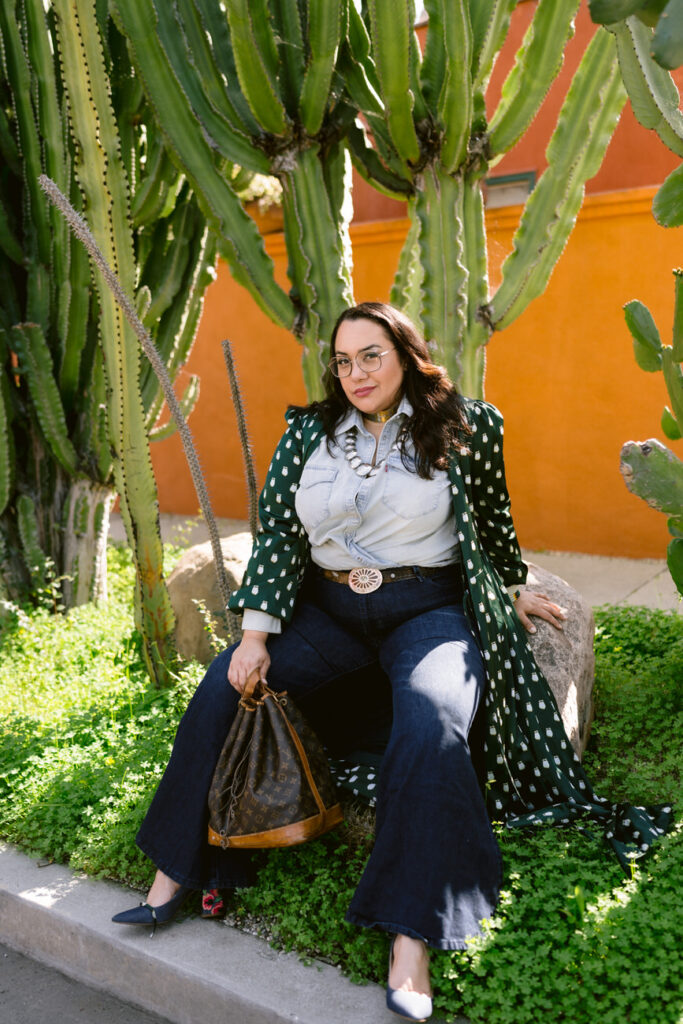 A person sitting comfortably among cacti, dressed in denim, with a green patterned kimono draped over, and a vintage designer bag in hand.