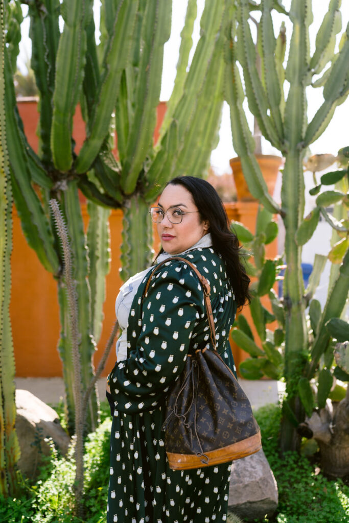 A rear view of a person walking away, dressed in a green patterned long coat and denim, with a vintage designer handbag on the shoulder, with large cacti in the background.