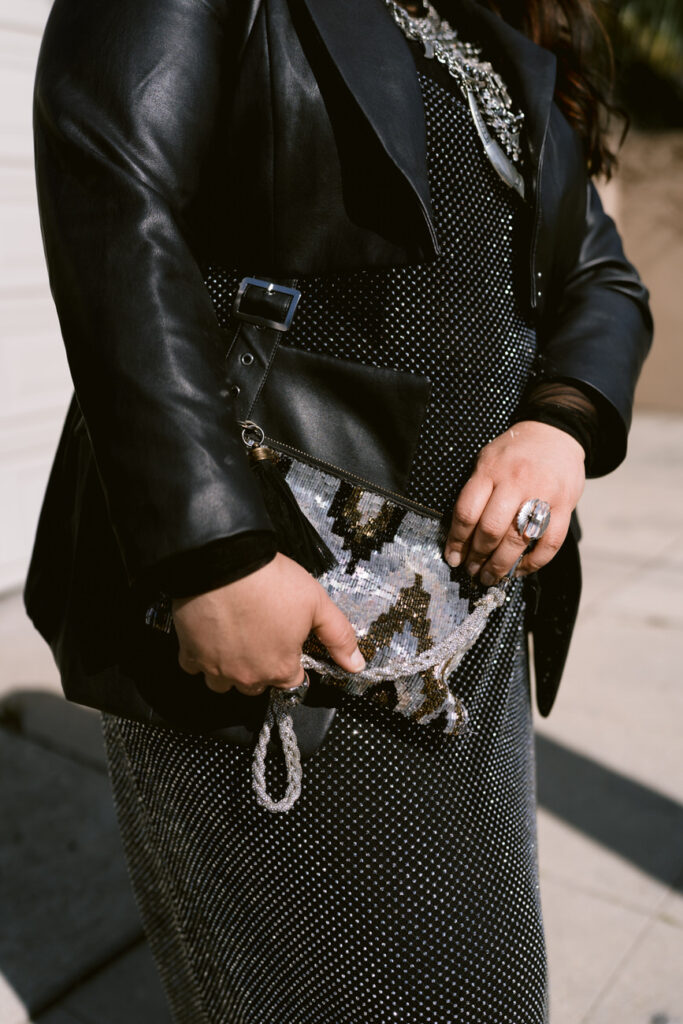 Close-up of a person's torso showing a layered chunky necklace, a black leather jacket, and a sparkling silver skirt, with one hand holding a sequined clutch.