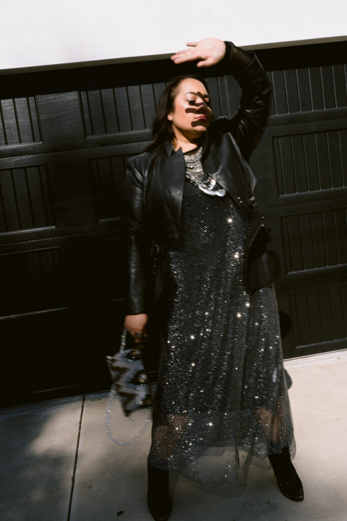 Dynamic photo of a person in motion, wearing a black leather jacket and sparkling skirt, one arm raised, creating a playful and carefree vibe.