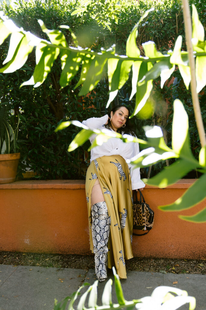 A person standing in a garden, partially obscured by foliage, dressed in a white blouse, mustard skirt with zebra details, and snake print boots, holding a leopard print bag.