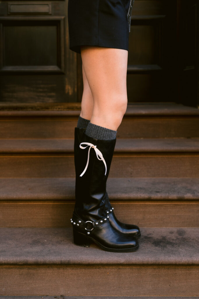 Black knee-high boots with a decorative white bow and silver buckle detail worn with dark shorts and grey socks against a backdrop of brownstone steps.