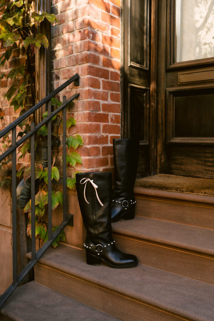 Black leather boots with a white bow detail stand alone on brownstone steps, complementing the green ivy and the rustic charm of the West Village, NYC.