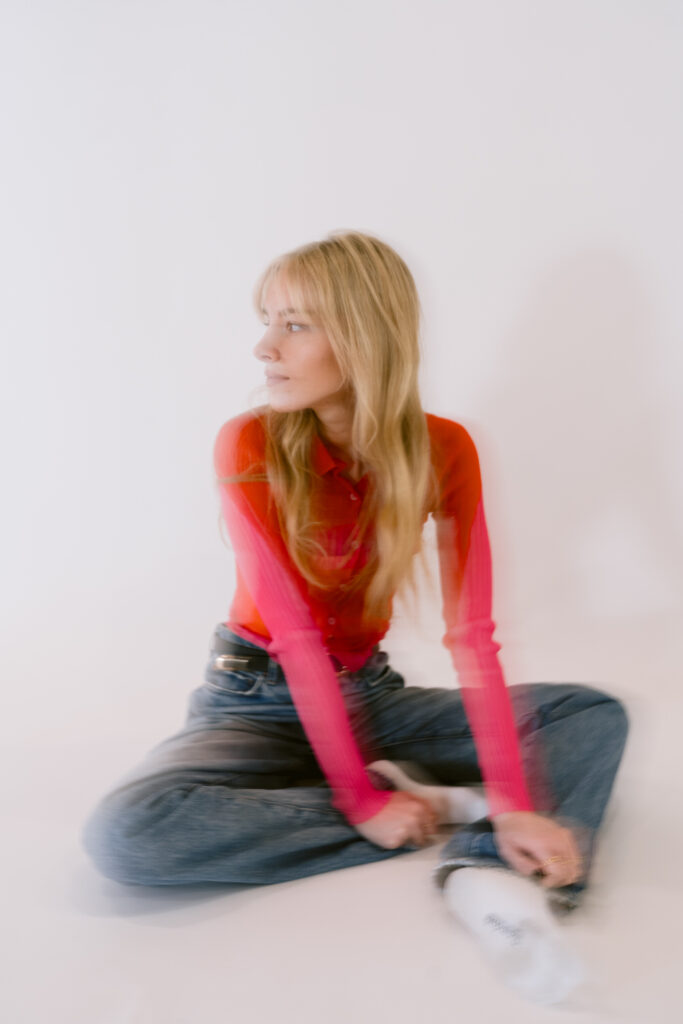 A softly focused photograph captures a young woman in motion, wearing a bright red top with pink sleeves and blue jeans, creating a blend of color and movement against a plain white background.