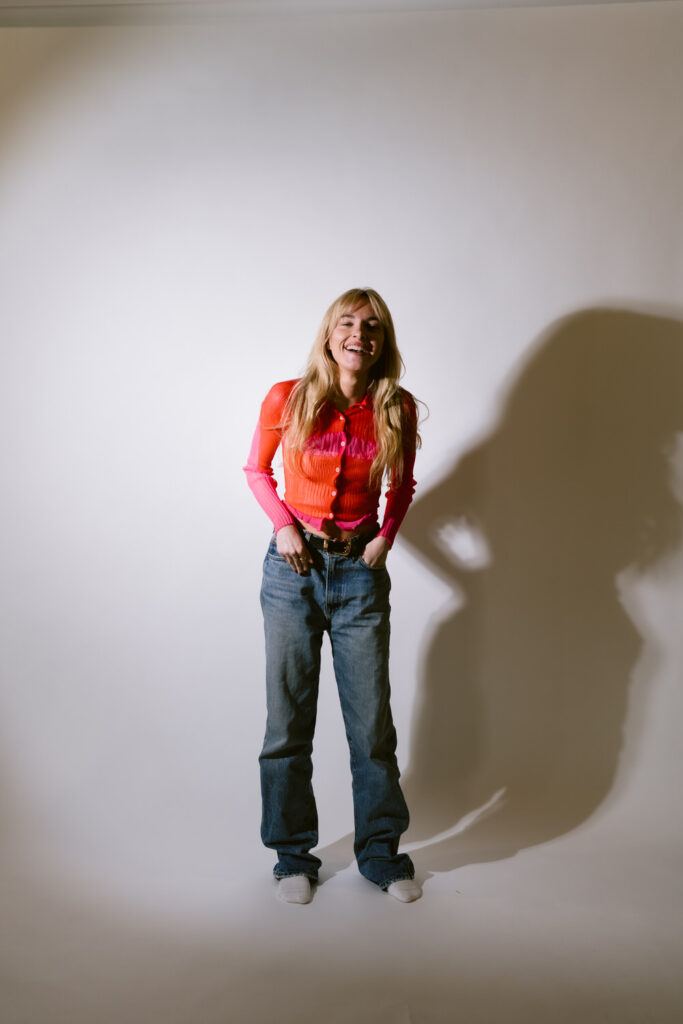 A happy person in a striking orange and pink long-sleeved mesh top paired with blue jeans stands laughing in front of a neutral backdrop, her shadow adding depth to the photo.